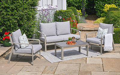 Capri Lounge Set with 2-seat sofa, 2 chairs and a coffee table, with grey cushions and a grey frame, on a stone patio in a garden