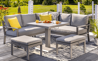 Image of Rio square corner set with adjustable table. Light grey cushioned square sofa, and 2 stools, surround a height adjustable table, on an black and white print rug topped patio with countryside in background.