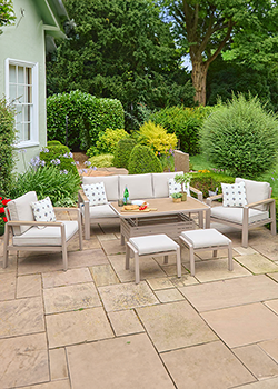 Metal frame lounge patio set, with cream cushions, glass topped rectangle coffee table. Trees in background.