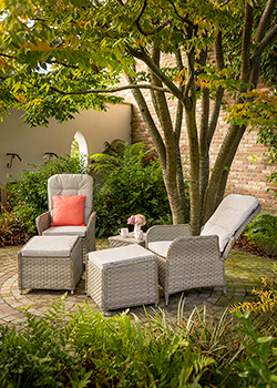 2 weave reclining chairs with cream cushions and yellow scatter cushions, and cushioned foot rests and side table, on a cobble floor with garden wall in background