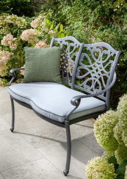 2 seater patterned metal bench in black with light grey cushion and green scatter cushion, on a patio with flowers in background