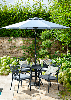 4 seat dining set with parasol, round table and 4 chairs, with a black patterned metal frame, grey/blue cushions and walled garden in background.