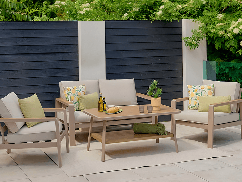 Sorrento Lounge Set with 2 seat sofa, 2 lounge chairs and a coffee table in a light brown colourway with cushions. Fence to background and trees overhead.